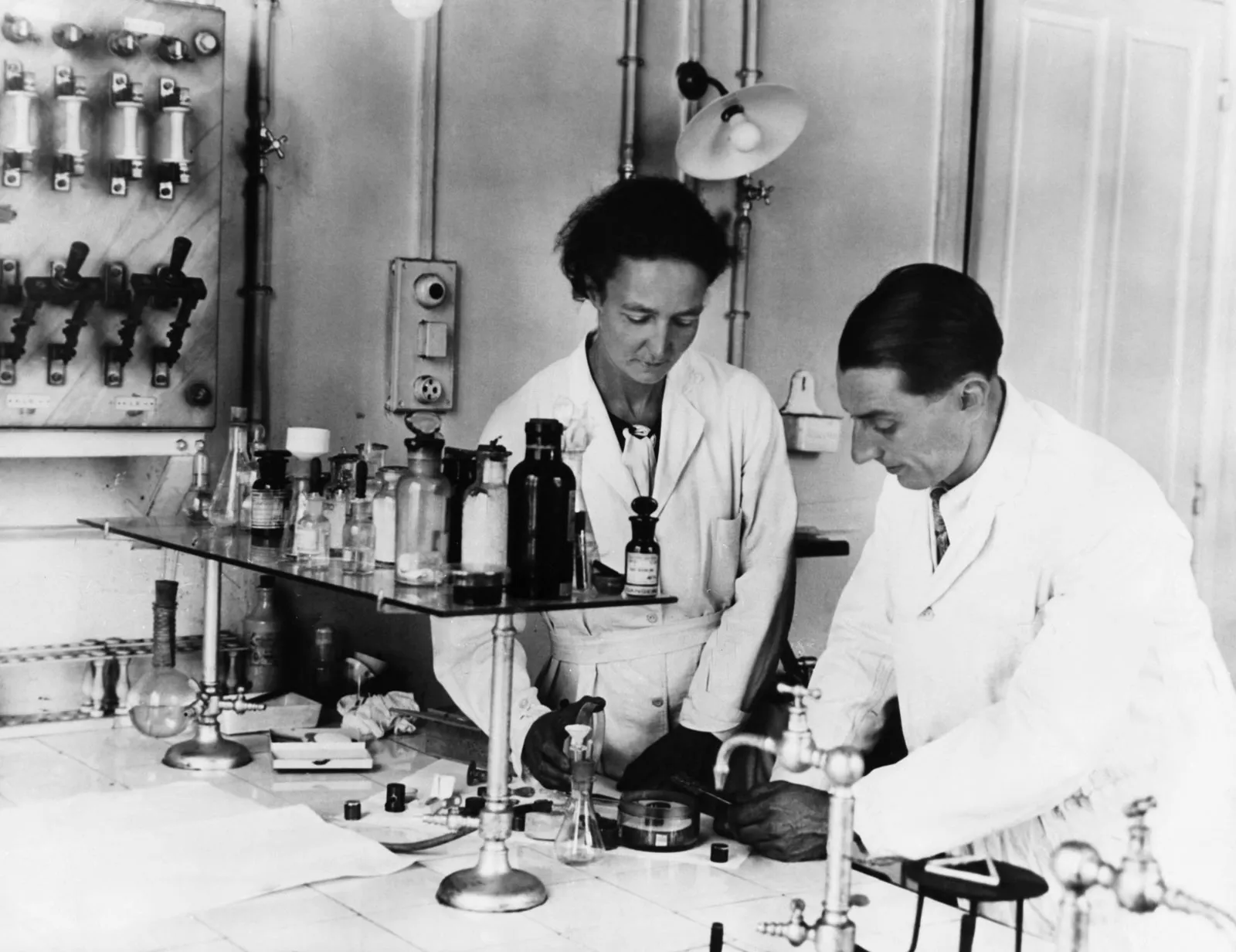 A black-and-white photograph of Irène and Frédéric Joliot-Curie working at a laboratory bench with glassware and an electrical switchboard behind them.